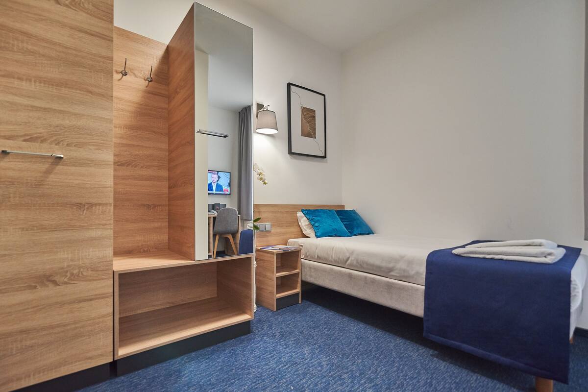 Una habitación de hotel moderna con un armario de madera, una cama con almohada azul, una mesita de noche pequeña y una lámpara fijada a la pared.
