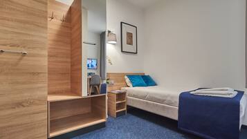 Una habitación de hotel moderna con un armario de madera, una cama con almohada azul, una mesita de noche pequeña y una lámpara fijada a la pared.