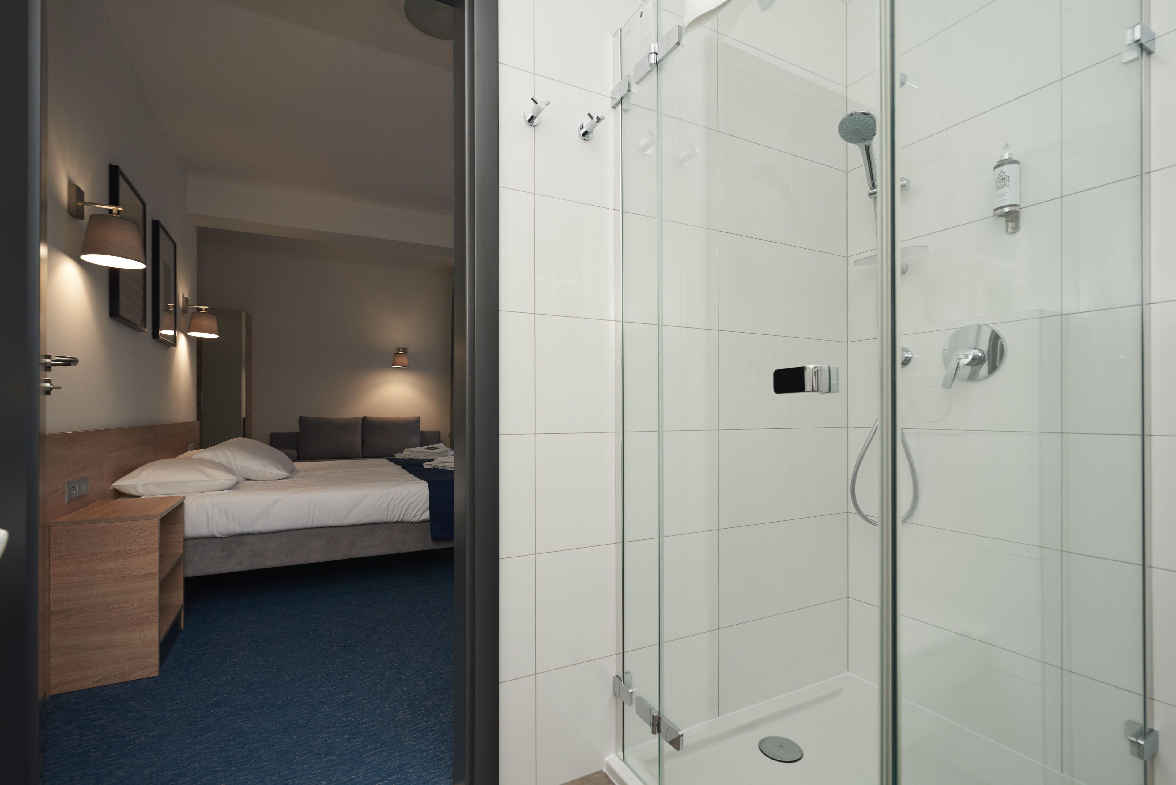 Habitación de hotel con cama, mesita de noche y zona de ducha con paredes de vidrio.