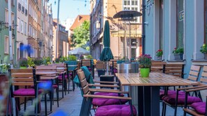 Um café ao ar livre com mesas e cadeiras de madeira, plantas em vasos e vista para a rua.