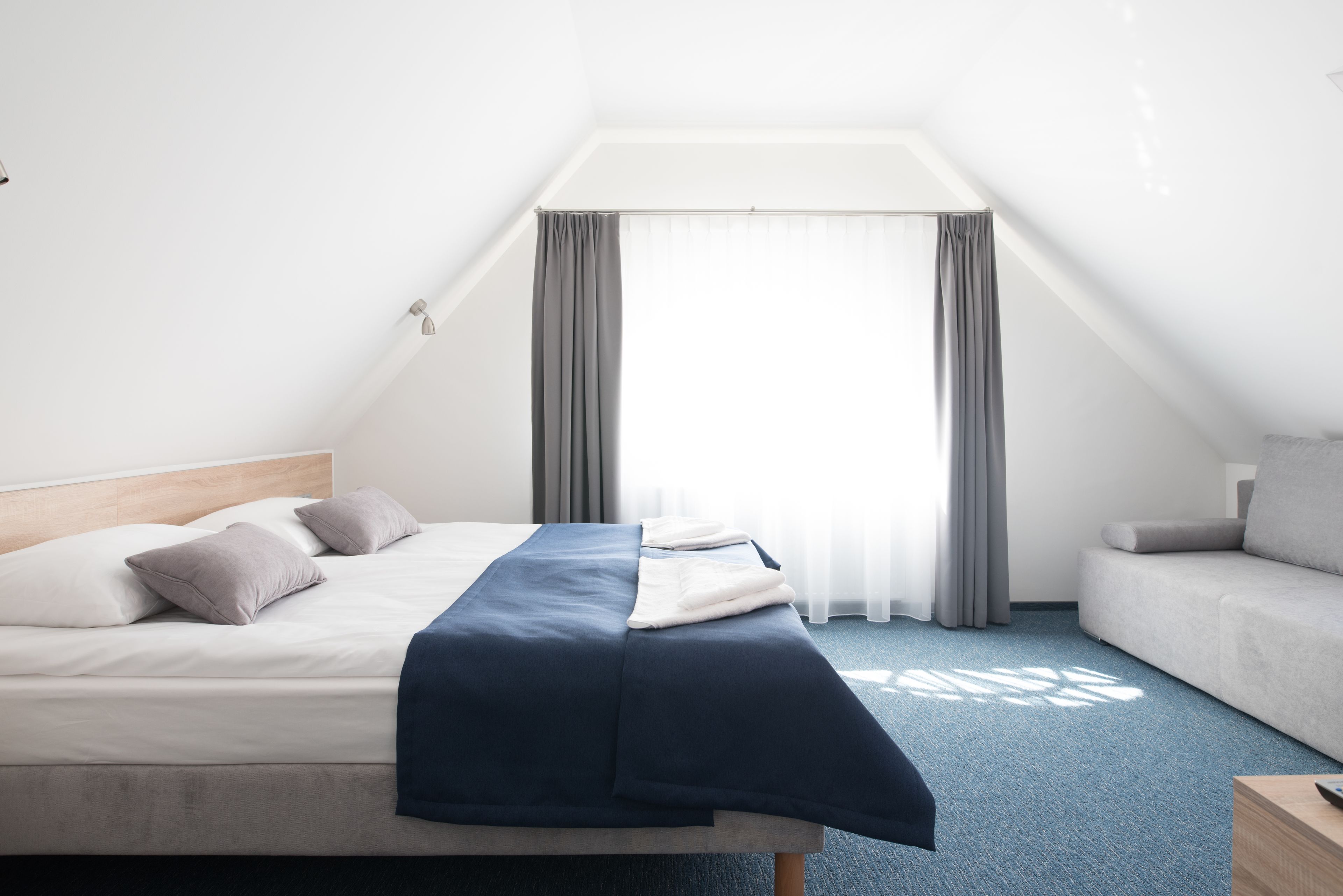 A neatly made bed with a blue blanket, a grey sofa, and a window with grey curtains.