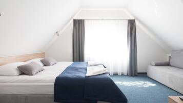 A neatly made bed with a blue blanket, a grey sofa, and a window with grey curtains.