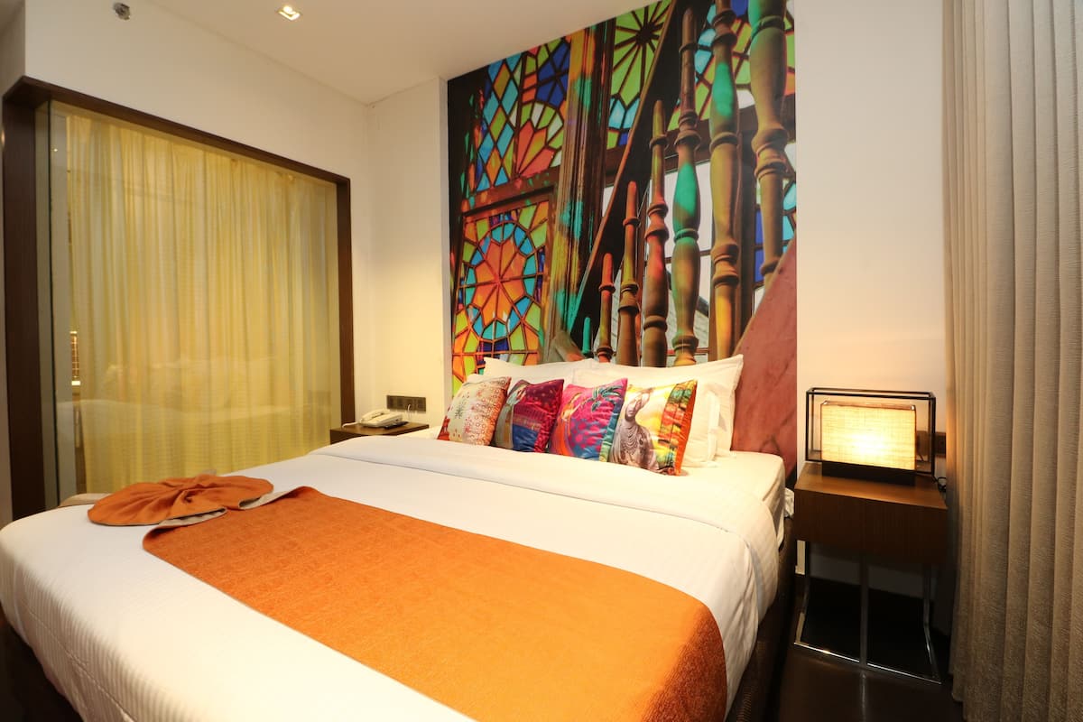 A hotel room with a large bed, a nightstand with a lamp, a window with curtains, and a colorful wall mural.