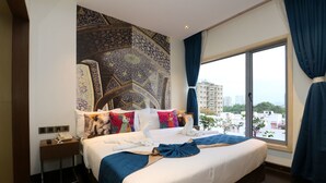 A hotel room with a large bed, a nightstand, a wall with intricate patterns, and a view of buildings through a large window. - Hotel Gallery 67 (Kolkata)