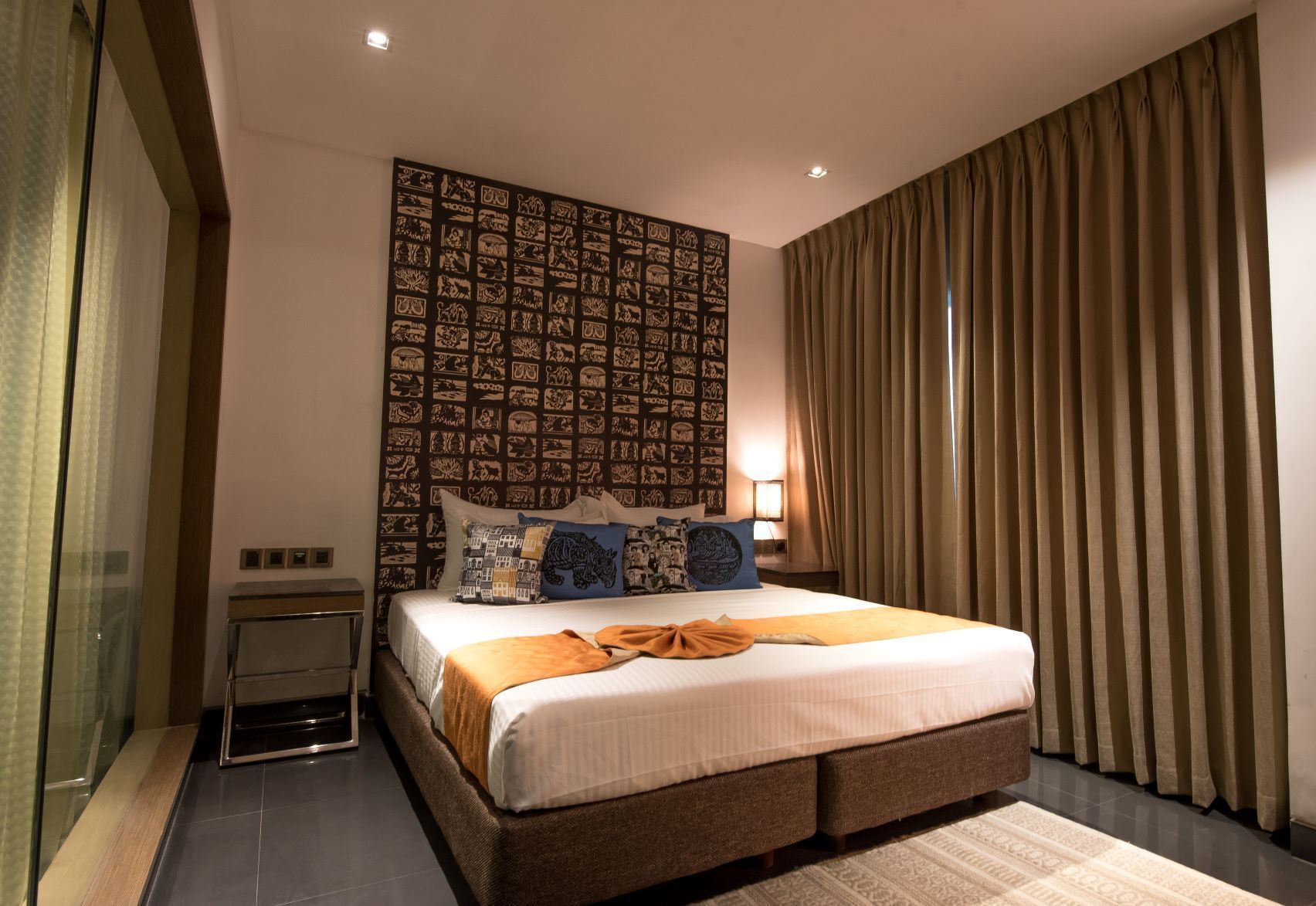 A modern hotel room with a large bed, a patterned headboard, and a view of the city through floor-to-ceiling windows.