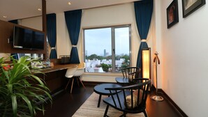 A modern hotel room with a large window, a flat-screen TV, a dining area with chairs, a small table, and a potted plant. - Hotel Gallery 67 (Kolkata)