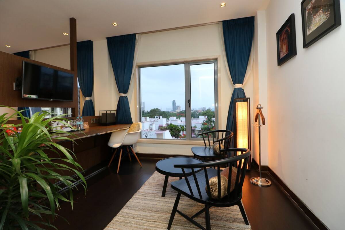 A modern hotel room with a large window, a flat-screen TV, a dining area with chairs, a small table, and a potted plant.