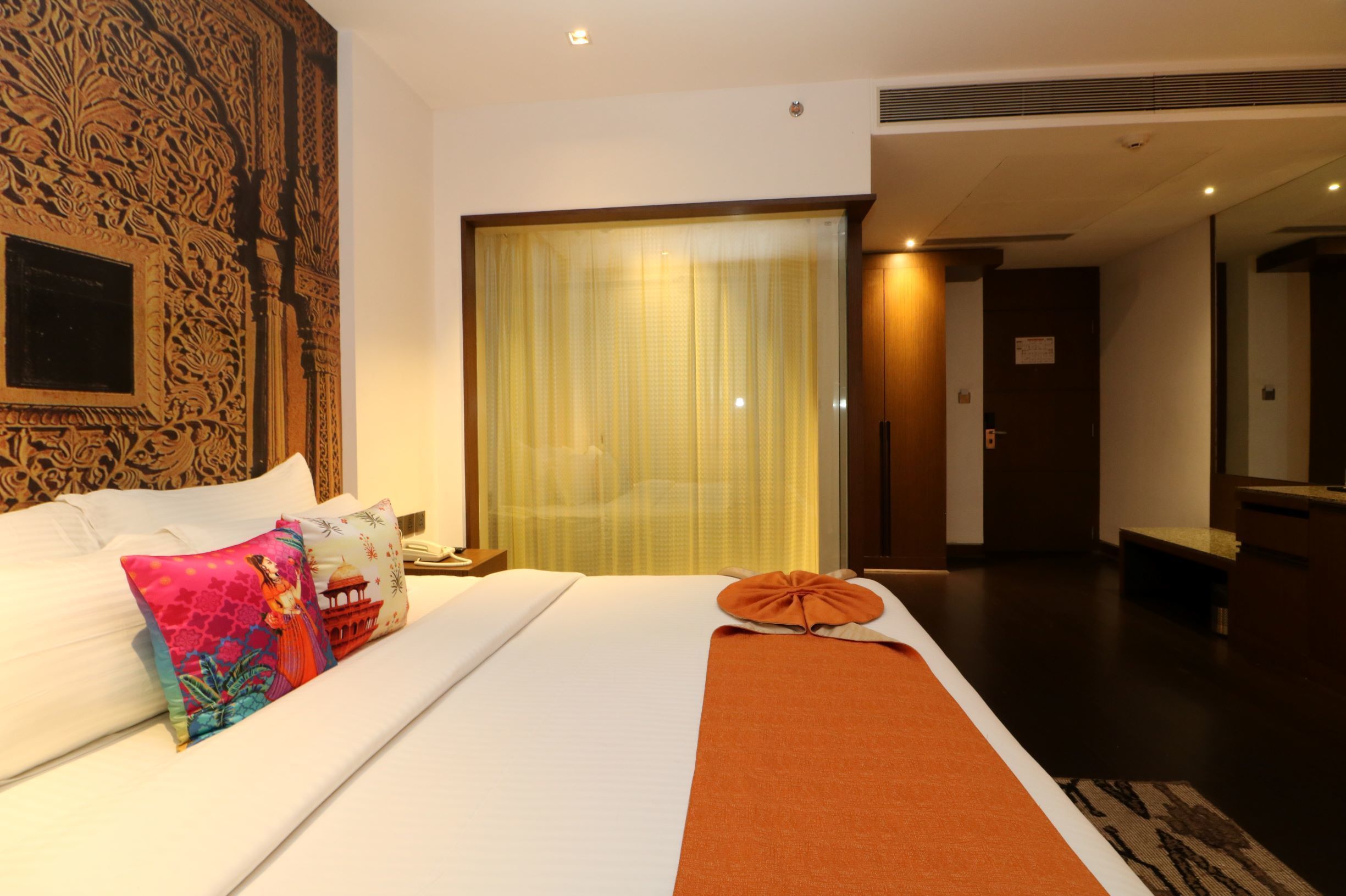 A hotel room with a large bed, decorative pillows, a large window with curtains, and a wooden floor.