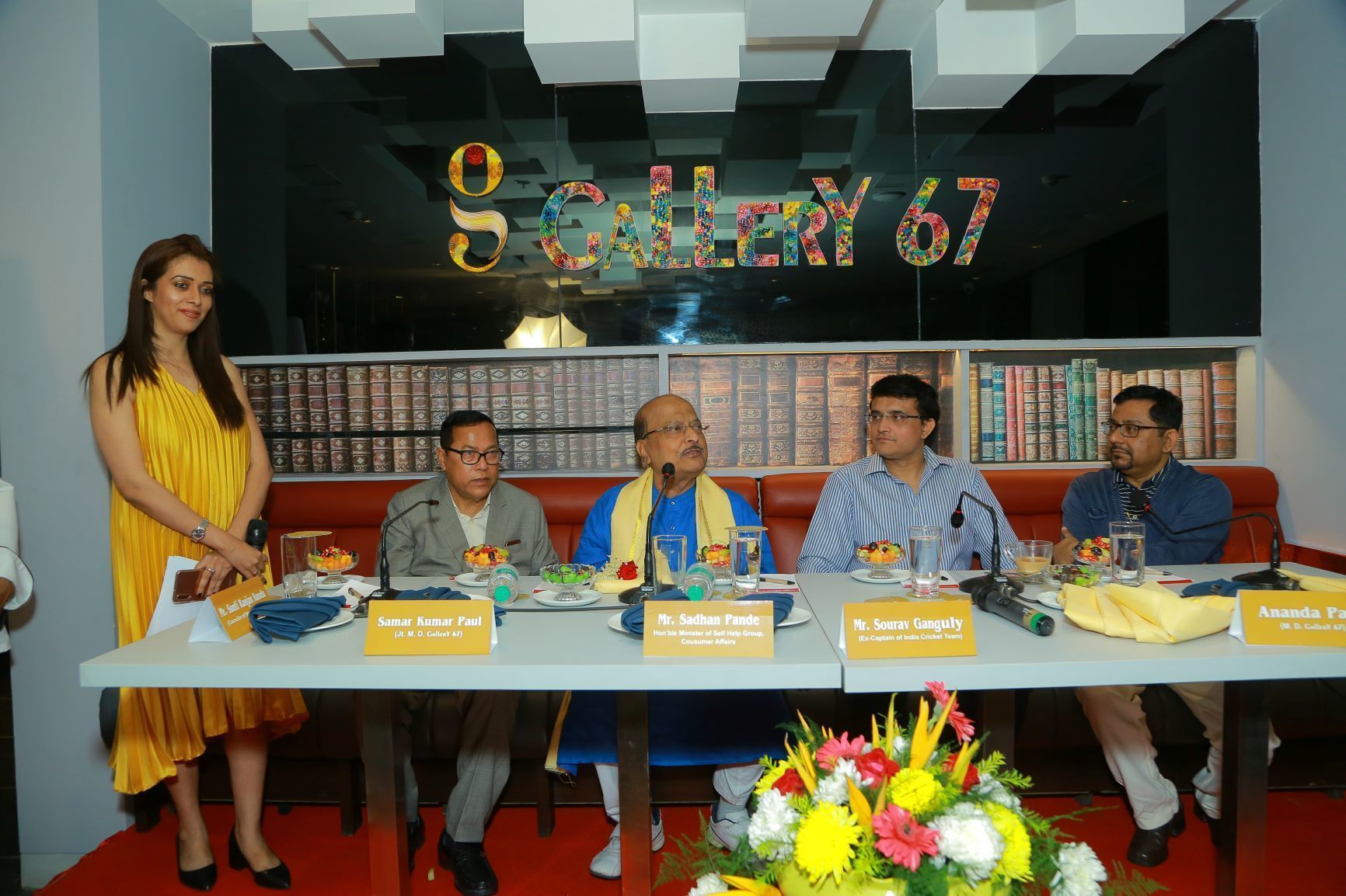 A panel discussion with five individuals seated at a table with nameplates, in a room with bookshelves in the background.
