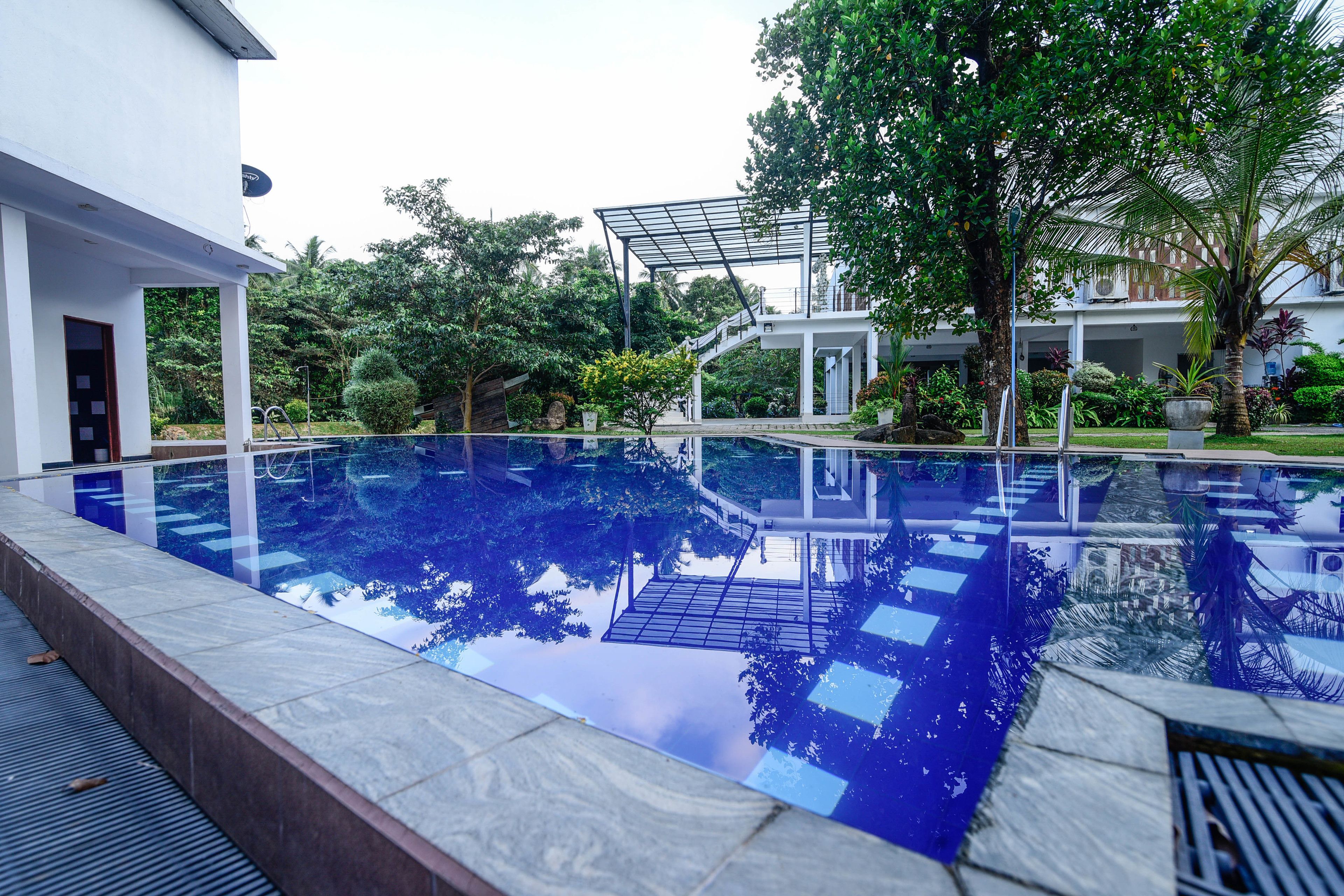 Una piscina con fondo de cristal, rodeada de vegetación y un edificio blanco.