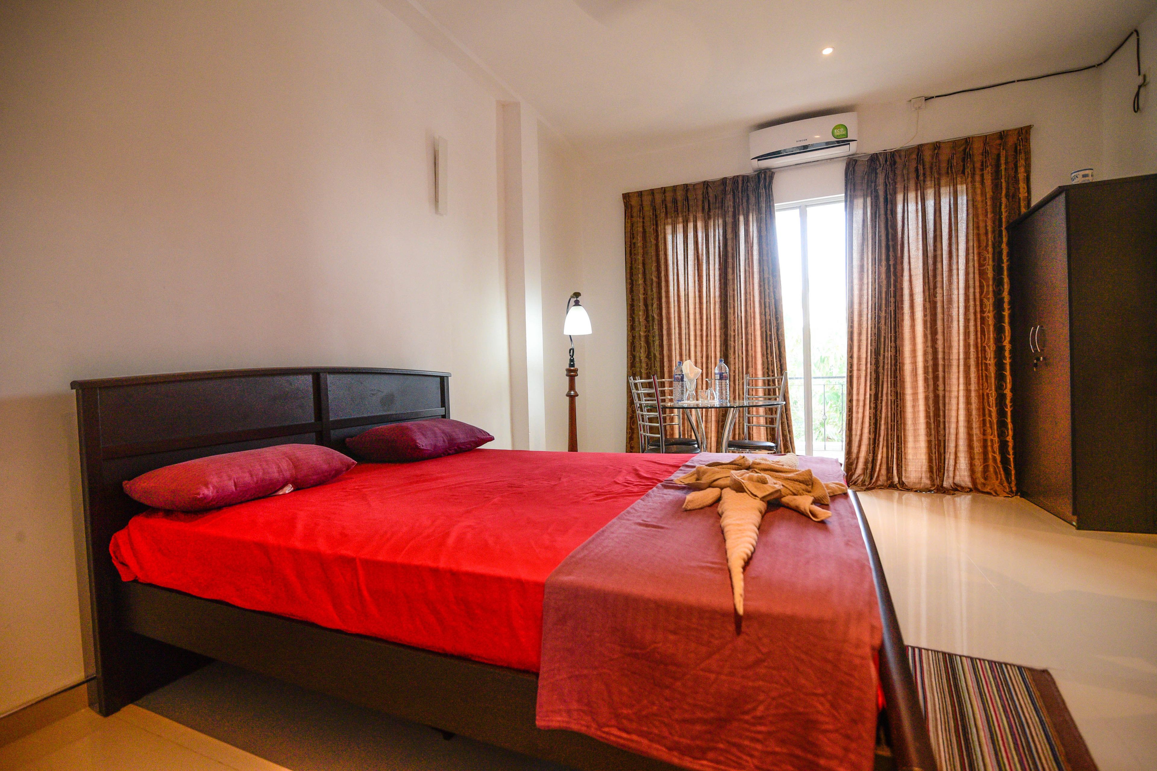 A bedroom with a bed, a nightstand, a lamp, a window with curtains, and an air conditioning unit.