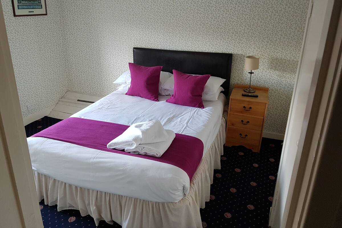 A neatly made bed with white and purple bedding, a wooden nightstand with a lamp, and a patterned carpet.