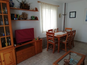 A living room with a wooden TV stand, a dining table with chairs, a glass coffee table, and a bookshelf.