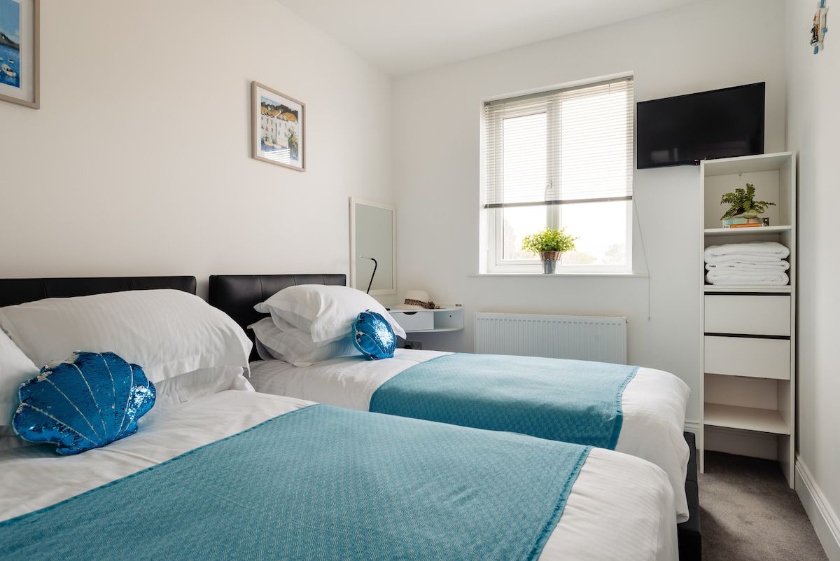 A hotel room with two beds, a television, a window with blinds, a radiator, and a shelf with towels.