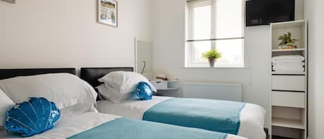 A hotel room with two beds, a television, a window with blinds, a radiator, and a shelf with towels.