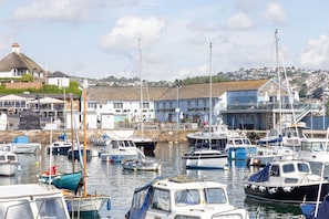A marina with various boats docked, a building labeled 'Harbour Light,' and a hillside in the background. - Quayside View (Paignton)