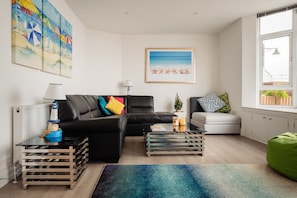 A living room with a black leather sofa, a white armchair, a coffee table, and a colorful rug. There are paintings on the wall and a window with a view. - Quayside View (Paignton)