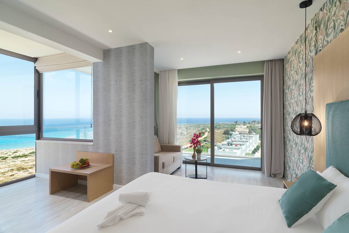 Una habitación de hotel moderna con una cama grande, una pequeña zona de estar, vistas a la playa y un balcón con vistas a la ciudad.