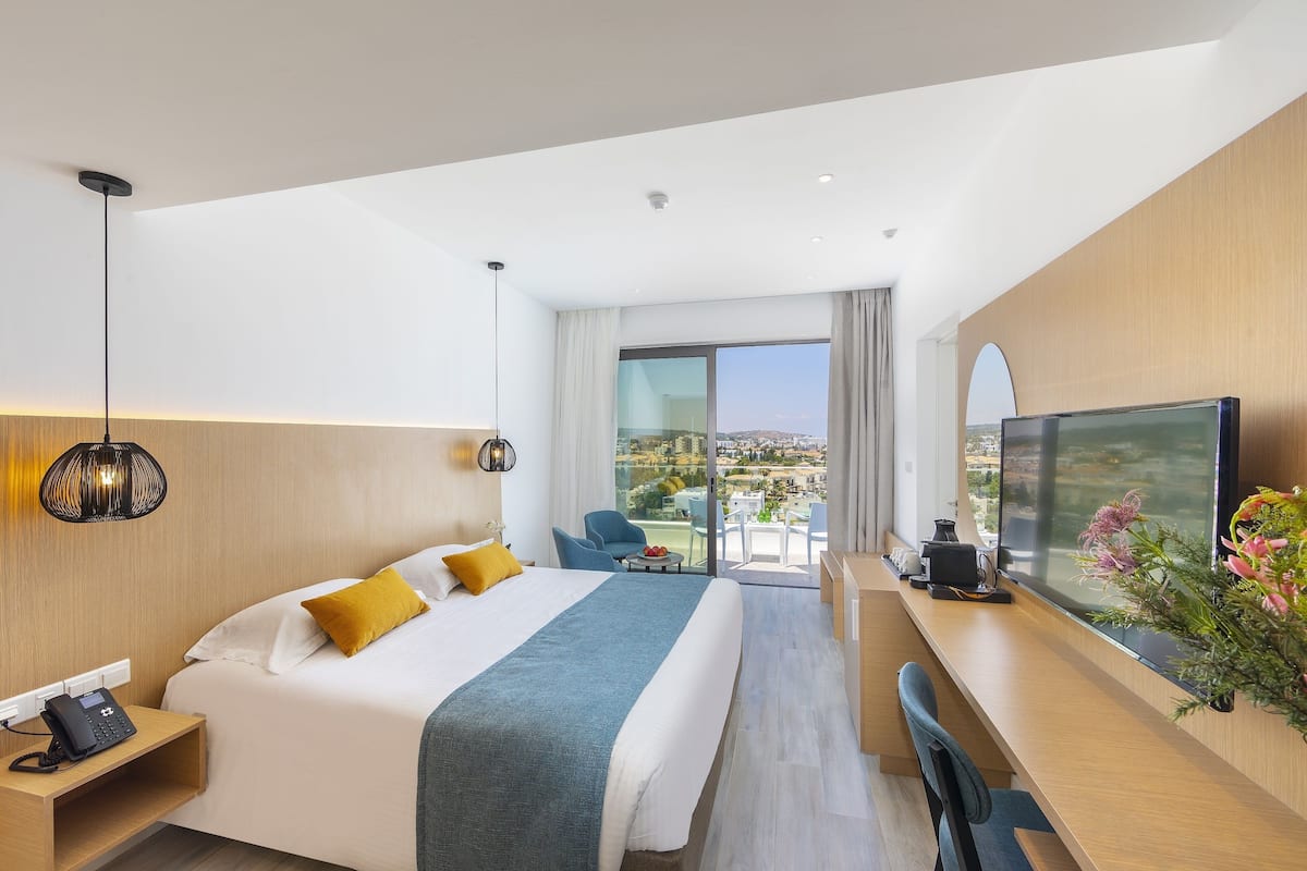 Una habitación de hotel moderna con una cama grande, un escritorio con un televisor de pantalla plana, un teléfono y vistas de la ciudad a través de ventanas amplias.