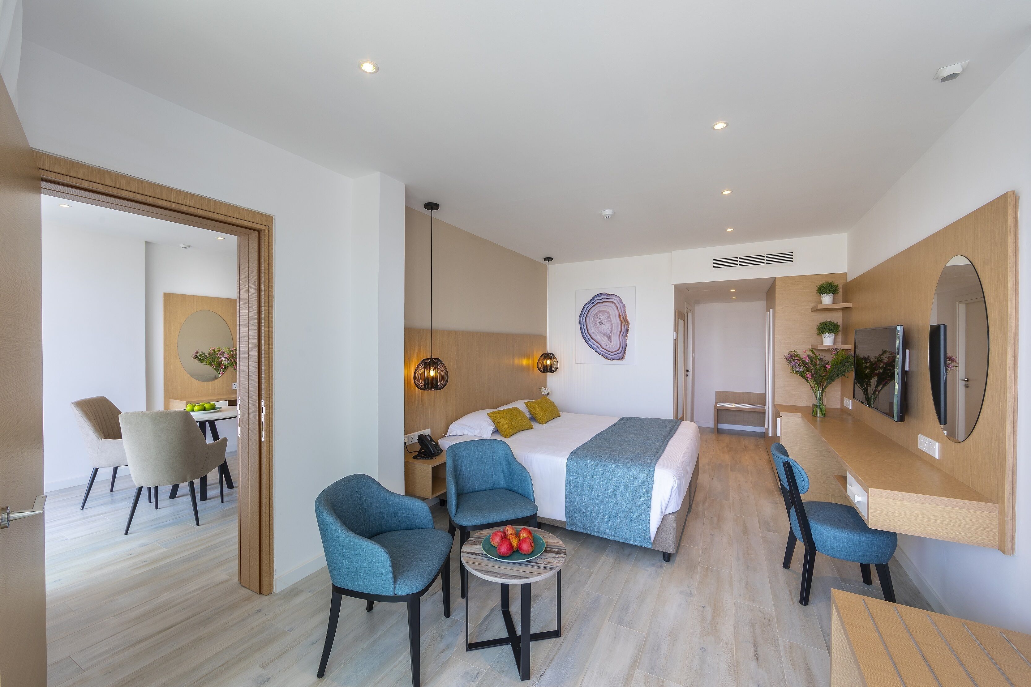 A modern hotel room with a bed, two blue armchairs, a small table with fruit, a desk with a computer, and a TV.