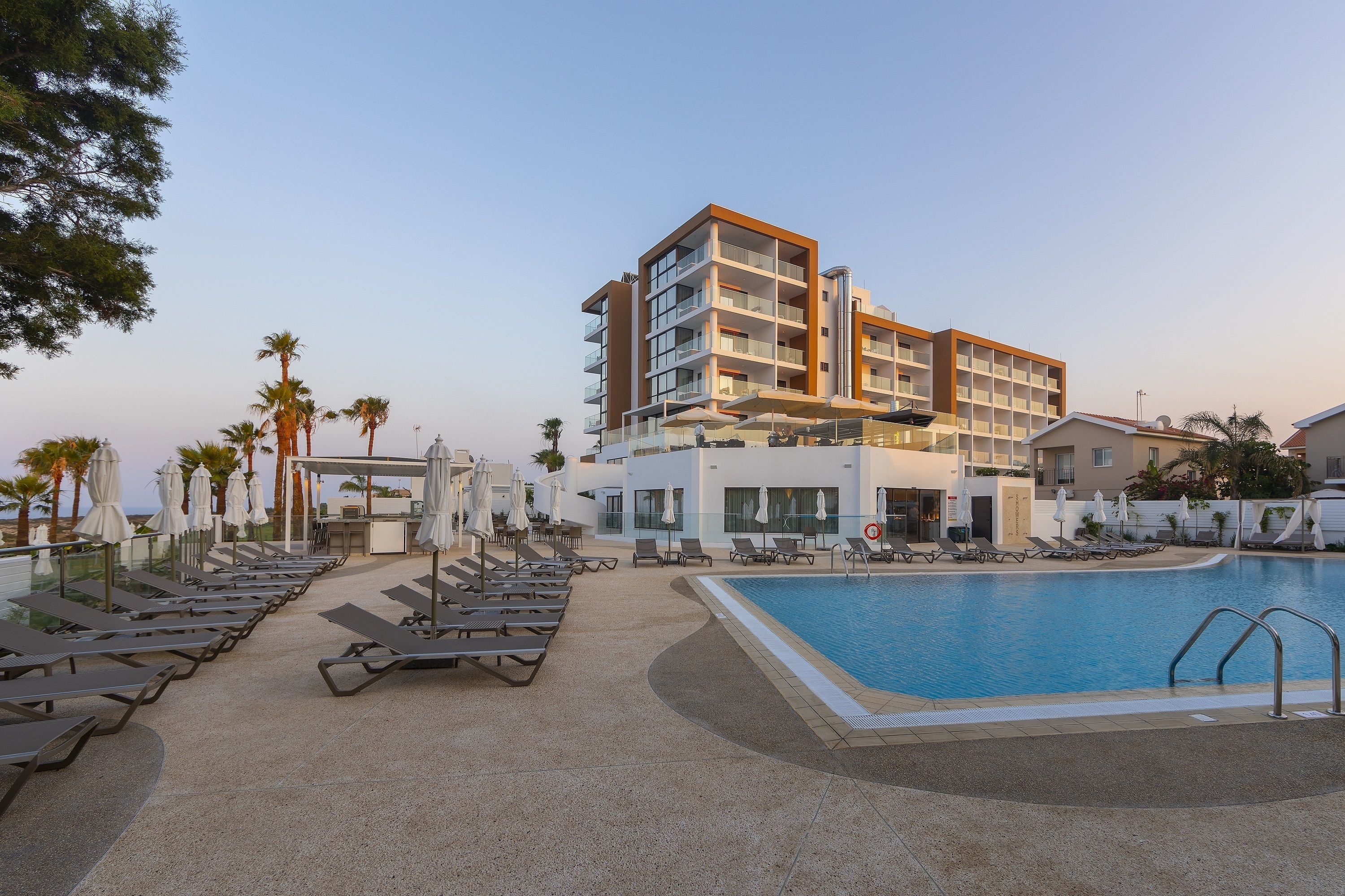 A poolside area with sun loungers and umbrellas, a modern building with multiple floors, and a clear blue sky.