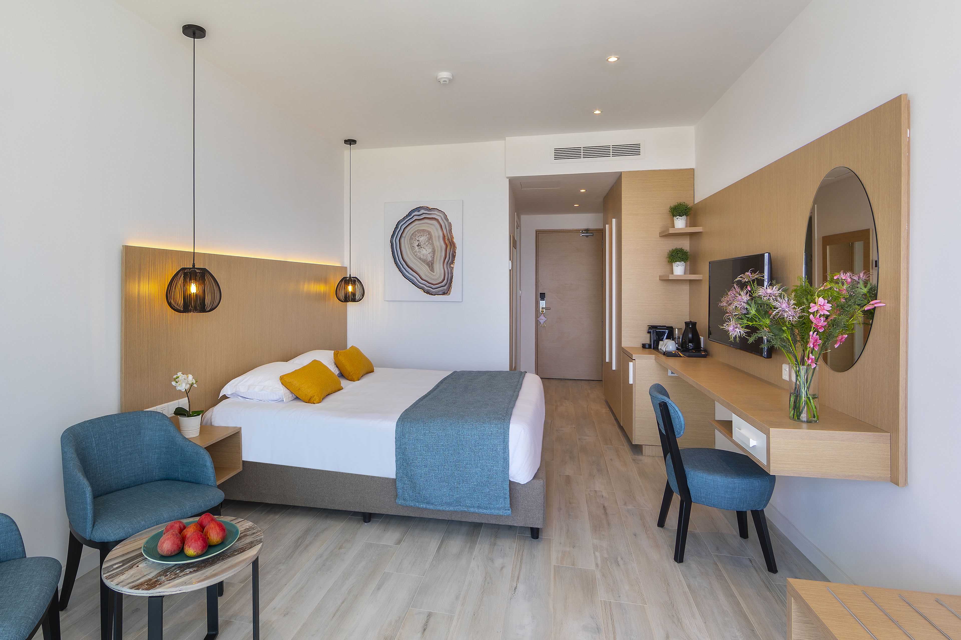 A modern hotel room with a bed, a desk, a chair, a small table with fruit, and a vase with flowers.
