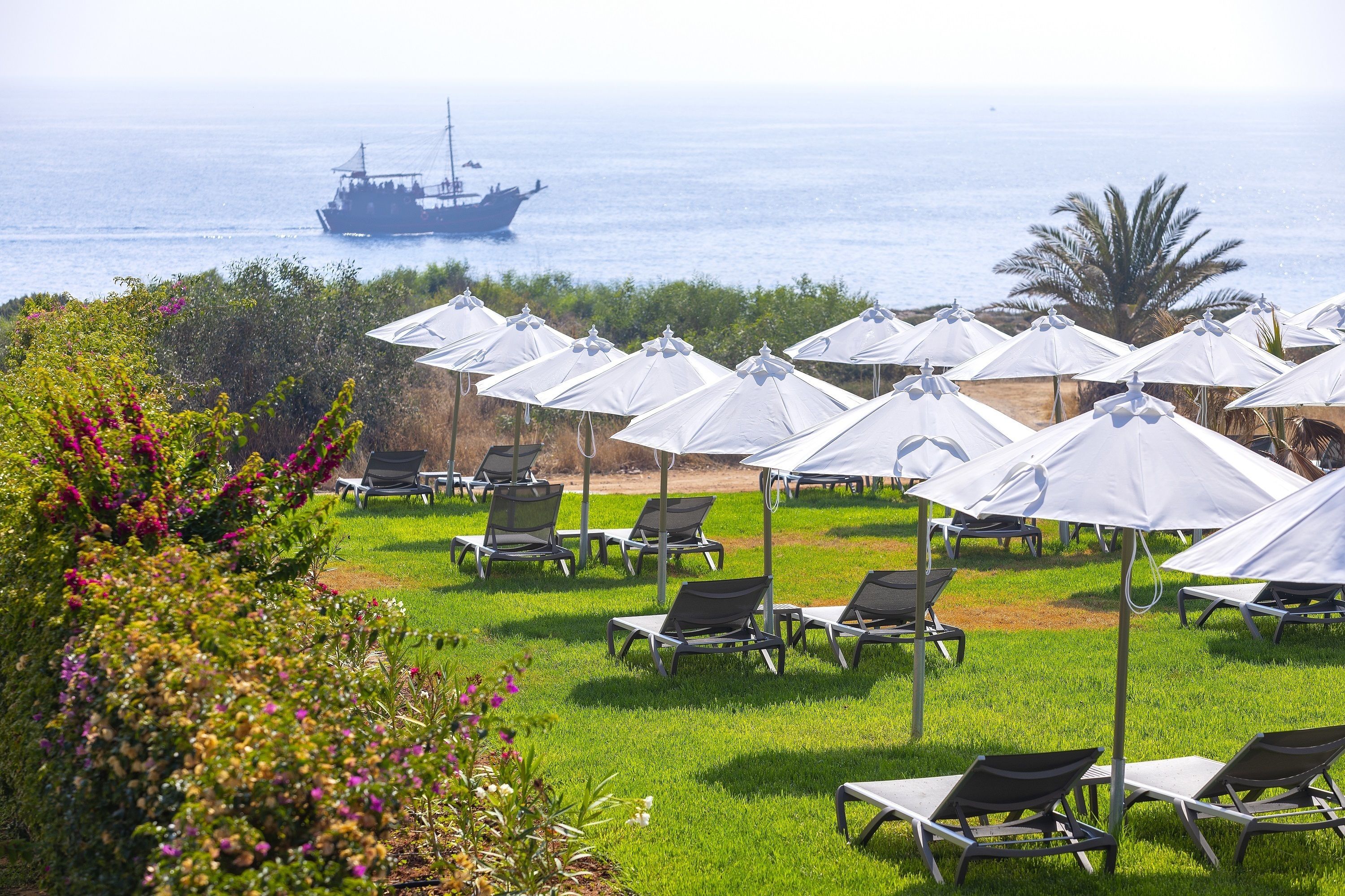 A coastal resort with rows of sun loungers under white umbrellas, a ship in the background, and a view of the sea.
