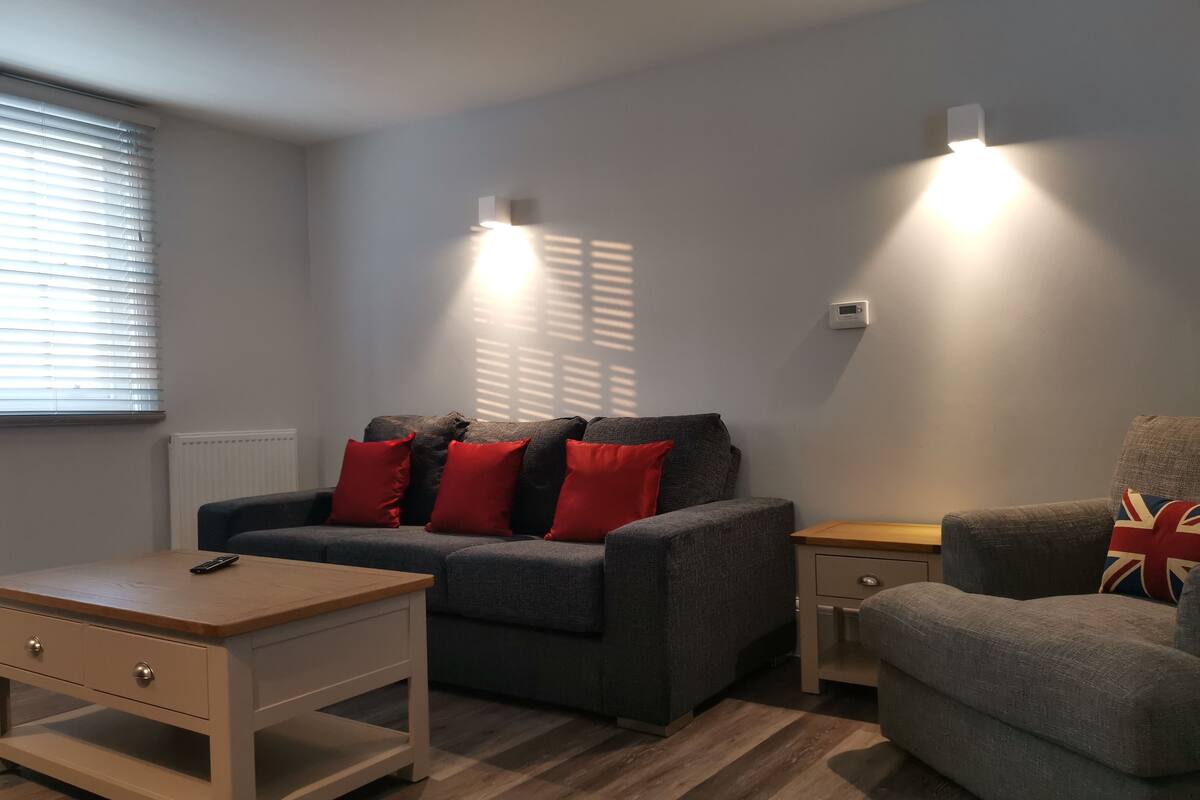 Un salon moderne avec un canapé gris, des coussins rouges, une table basse en bois et un fauteuil gris agrémenté d’un coussin à motifs Union Jack.