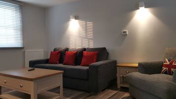 Un salon moderne avec un canapé gris, des coussins rouges, une table basse en bois et un fauteuil gris agrémenté d’un coussin à motifs Union Jack.