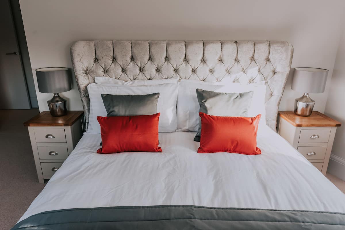 Un lit avec une tête de lit capitonnée, une literie blanche et deux coussins rouges. Il y a deux tables de chevet avec des lampes et une petite table en bois sur le côté.