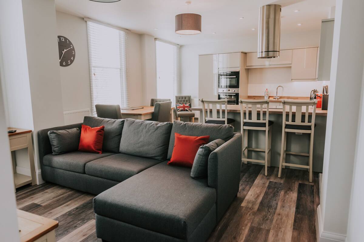 Un salon moderne avec un canapé d’angle gris, une table à manger en bois avec des chaises, un coin cuisine doté d’appareils électroménagers en acier inoxydable et une grande fenêtre avec des stores.