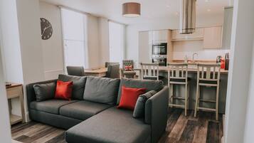 A modern living room with a grey sectional sofa, a wooden dining table with chairs, a kitchen area with stainless steel appliances, and a large window with blinds.