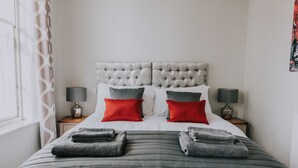 En pænt redt seng med en knapbetrukken gavl, to sengeborde med lamper, og et vindue med et geometrisk gardin.