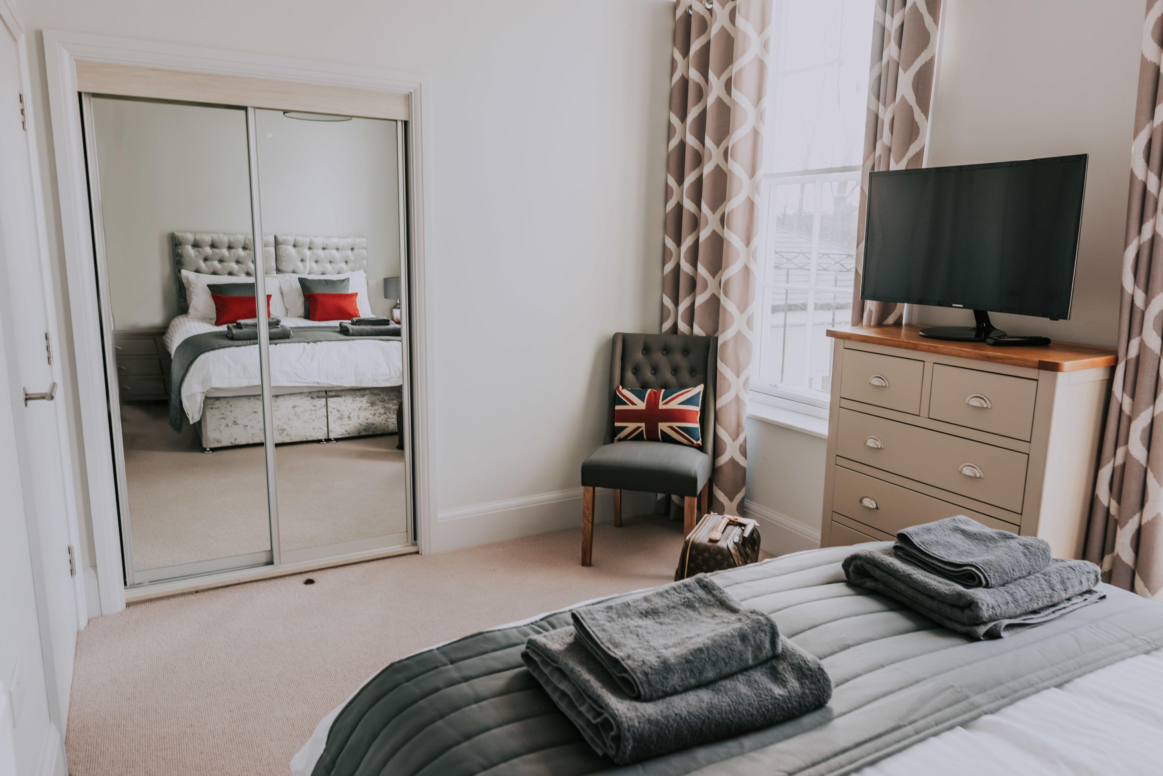 A bedroom with a bed, a chair, a TV, a wardrobe, and a window with curtains.