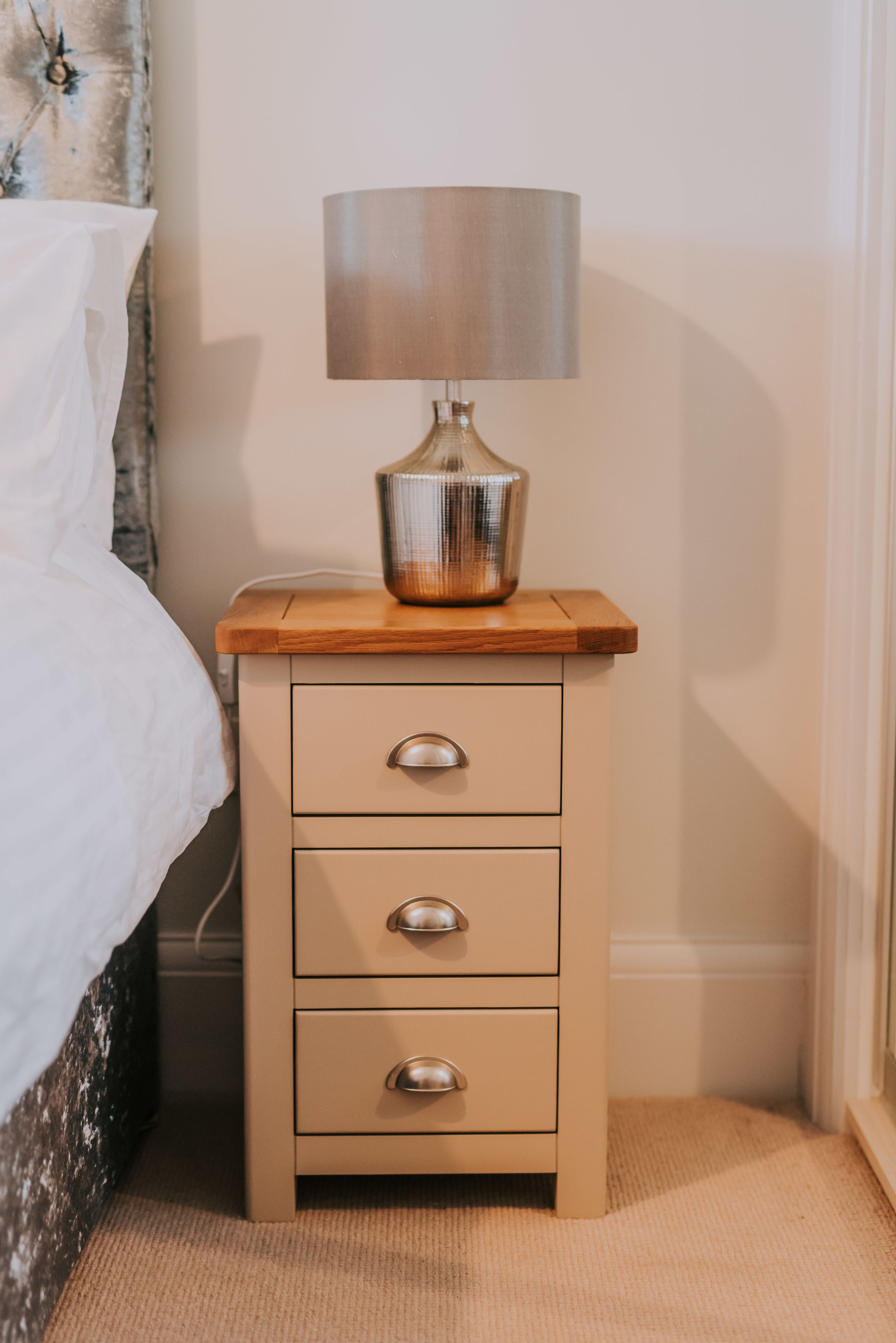 A bedside table with a lamp, a drawer, and a bed with a white bedspread.