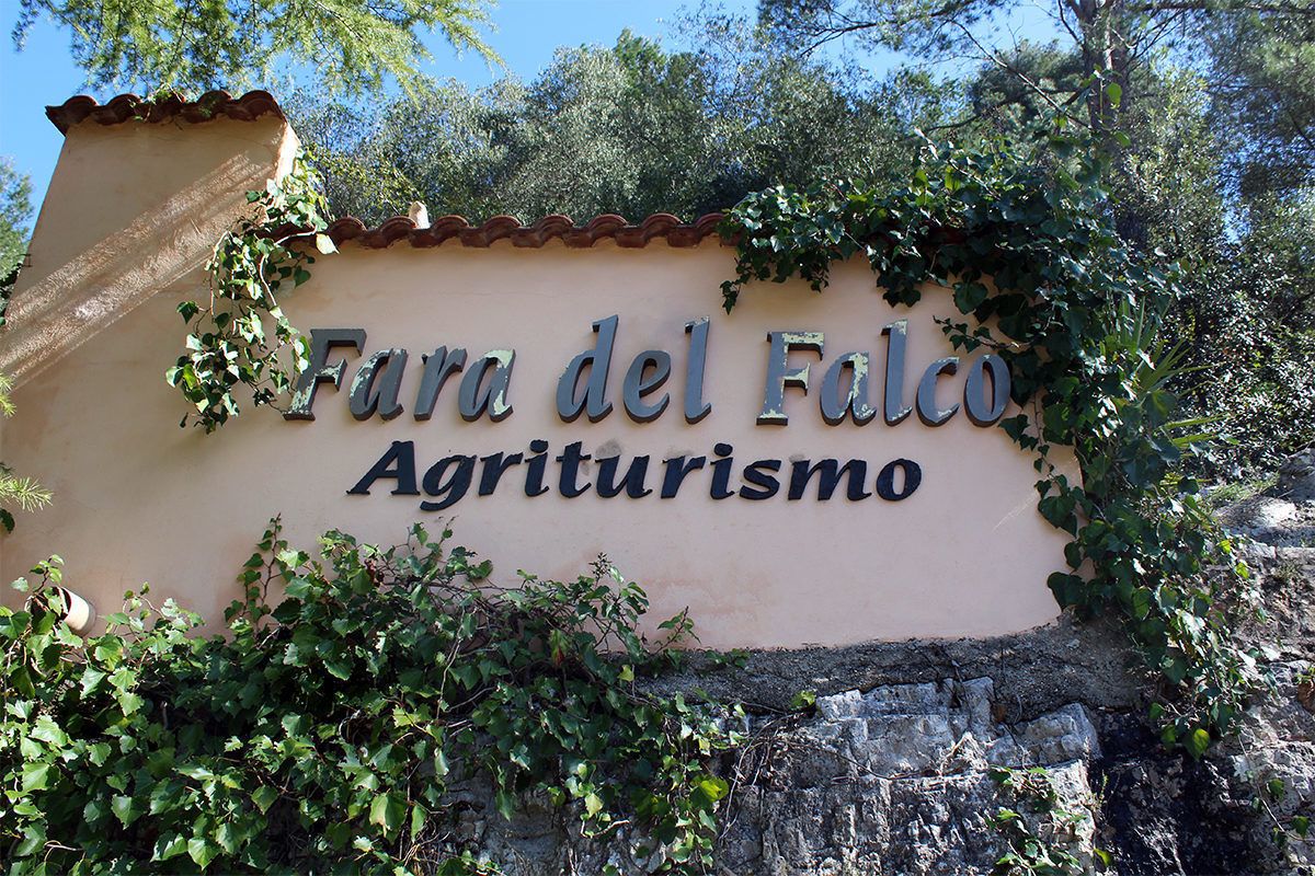 Naamplaatje voor 'Fara del Falco Agriturismo', met een rustieke stenen muur en begroeiing.