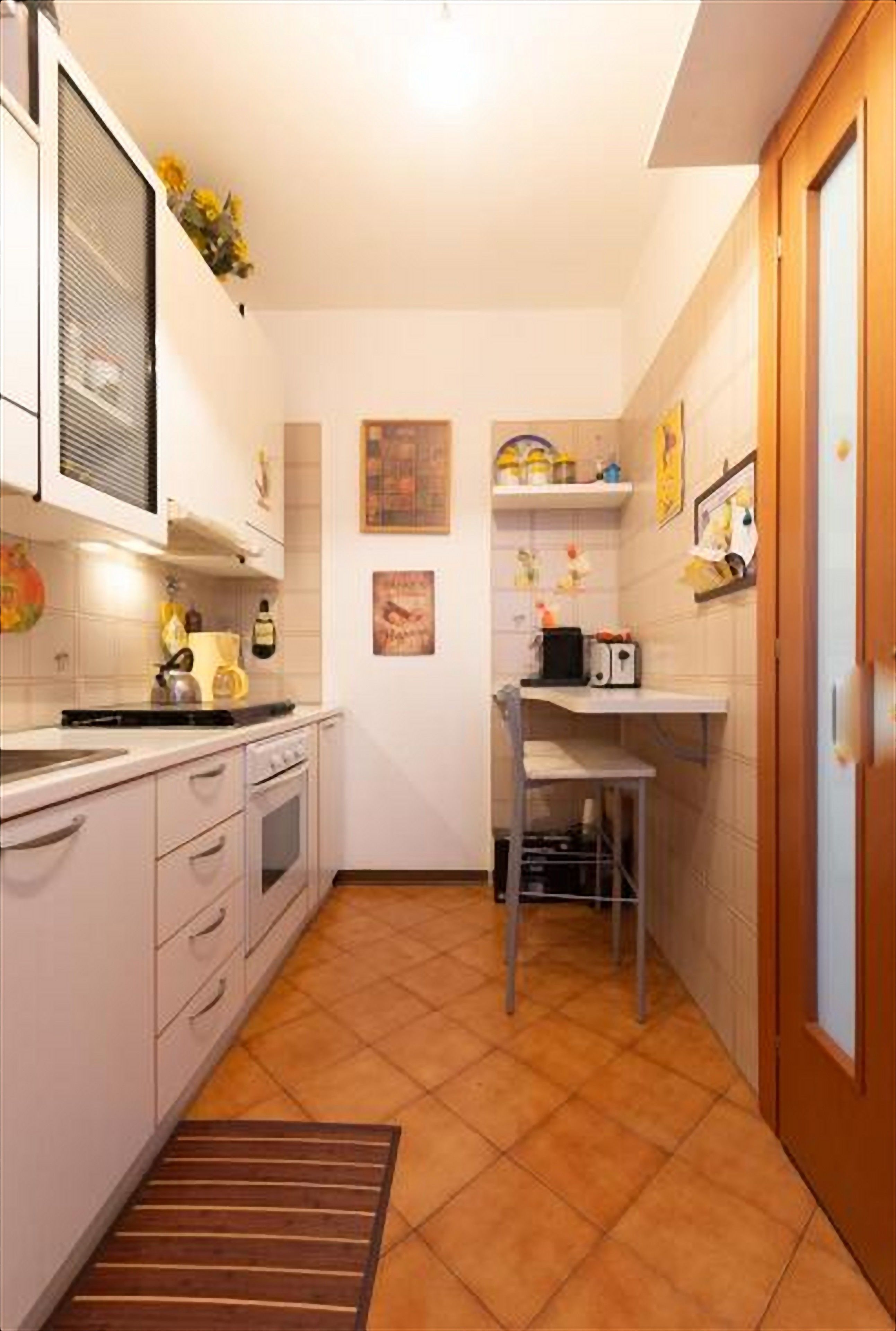 Una cocina compacta con armarios blancos, suelo de baldosas y un pequeño comedor con una silla y una mesa.