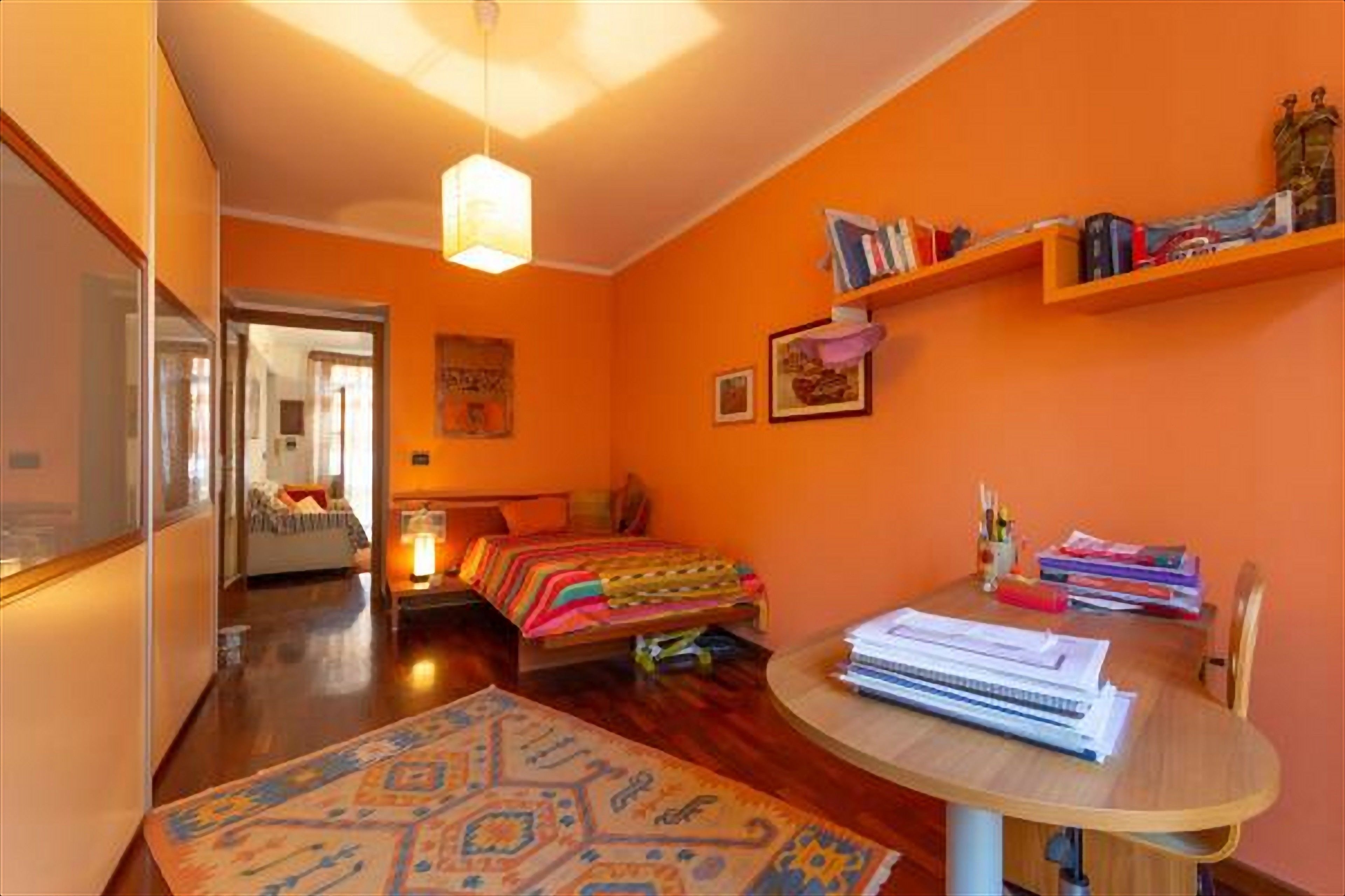 A room with an orange wall, a bed with a colorful blanket, a desk with papers, and a bookshelf with books and decorative items.
