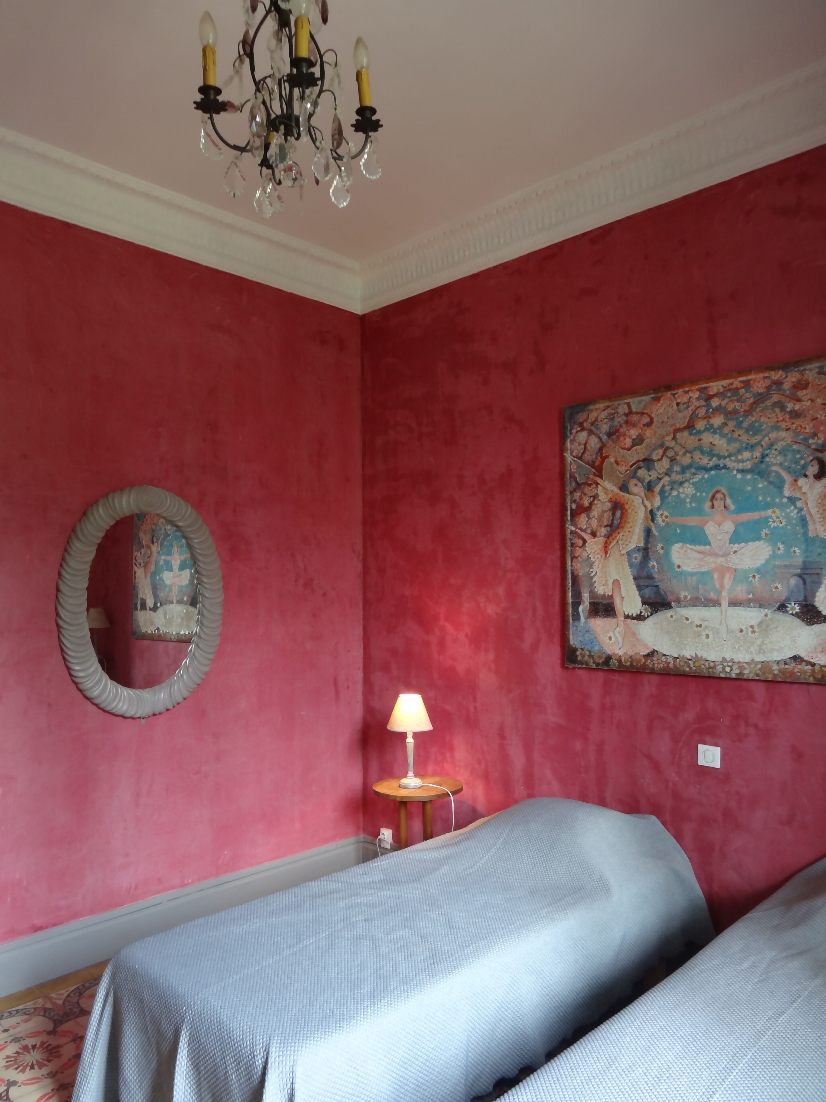 Een kamer met rode wanden, een kroonluchter, een ronde spiegel, een schilderij, een lamp en twee bedden.