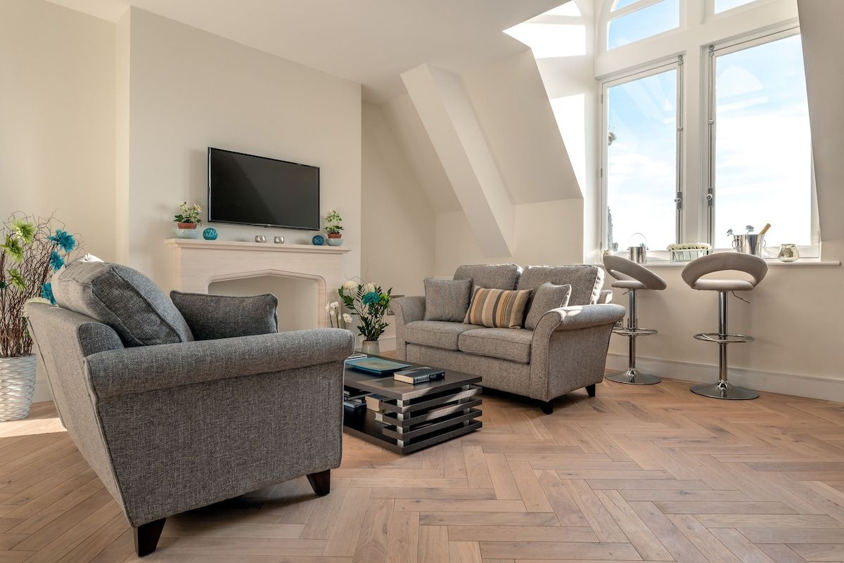 A modern living room with two grey sofas, a flat-screen TV, a fireplace, and a large window with a view.