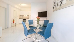 Un espace repas moderne avec une table en verre et des chaises bleues, une cuisine en arrière-plan et un téléviseur à écran plat fixé au mur.