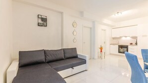 Un salon moderne avec un canapé d’angle gris, une petite table d’appoint sur laquelle est posé un vase, et un coin cuisine avec des meubles blancs et une cuisinière.