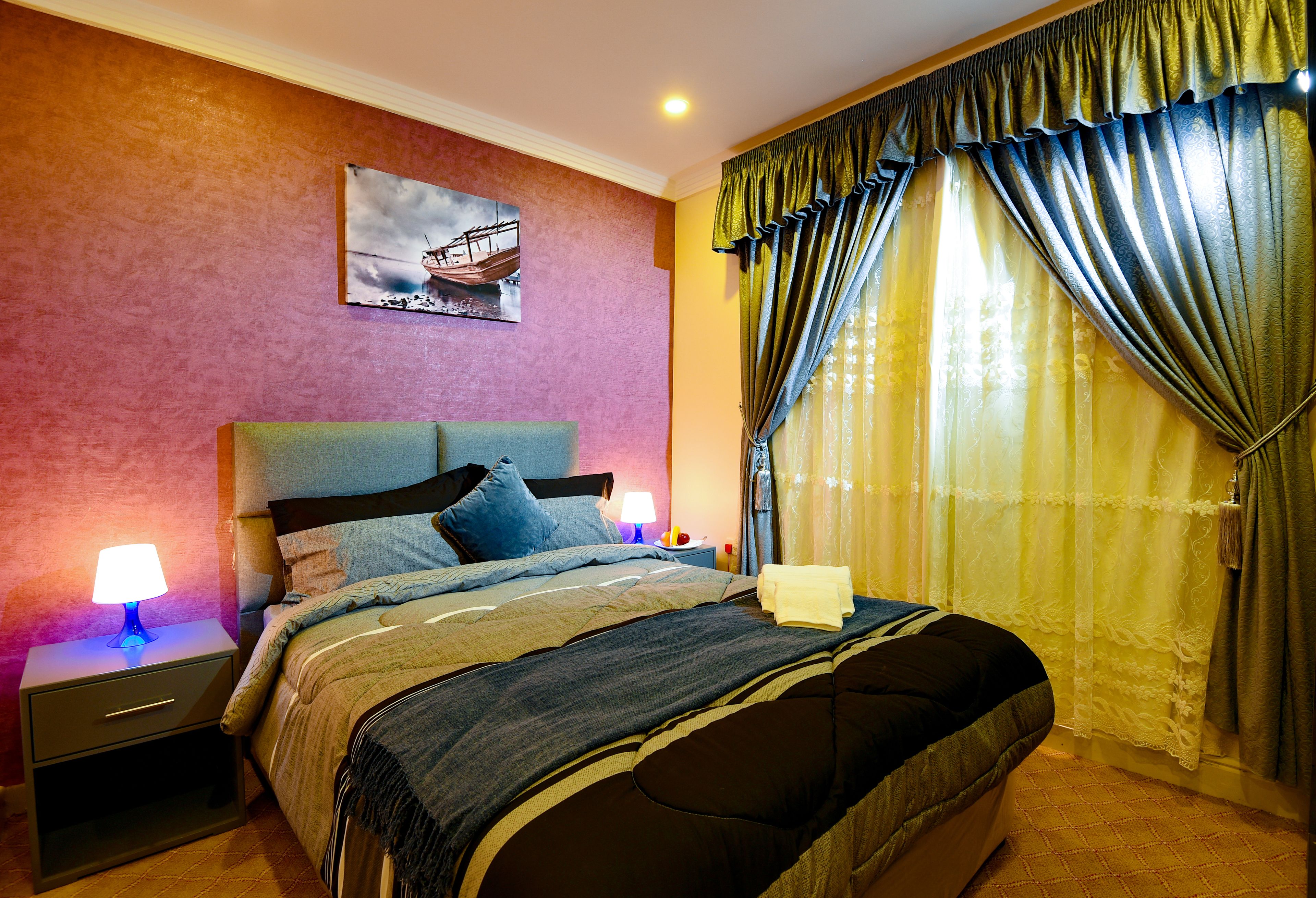A neatly made bed with a headboard, two bedside tables with lamps, a wall-mounted painting, and a window with curtains.