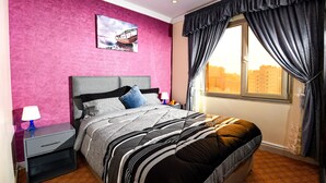 Un dormitorio con cama, mesita de noche, lámpara, ventana con cortinas y vista panorámica de la ciudad.
