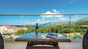 Un balcón con una mesa de vidrio, una botella y un libro, con vistas a un paisaje costero con casas y montañas.