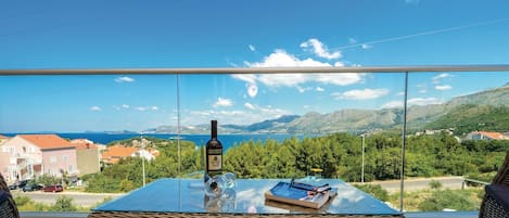 Een balkon met een glazen tafel, een fles en een boek, met uitzicht op een kustlandschap met huizen en bergen.
