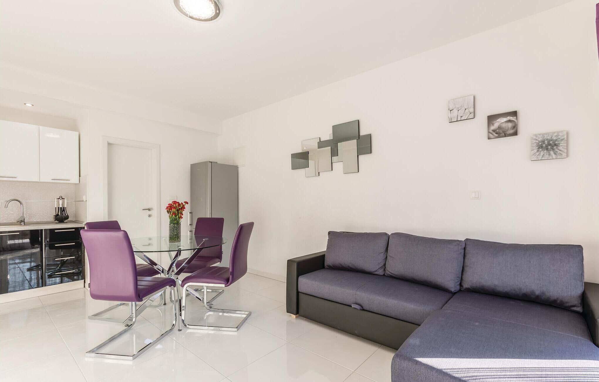 Een moderne woonkamer met een grijze bank en paarse stoelen, een glazen eettafel en een keuken met witte kasten en een spoelbak.
