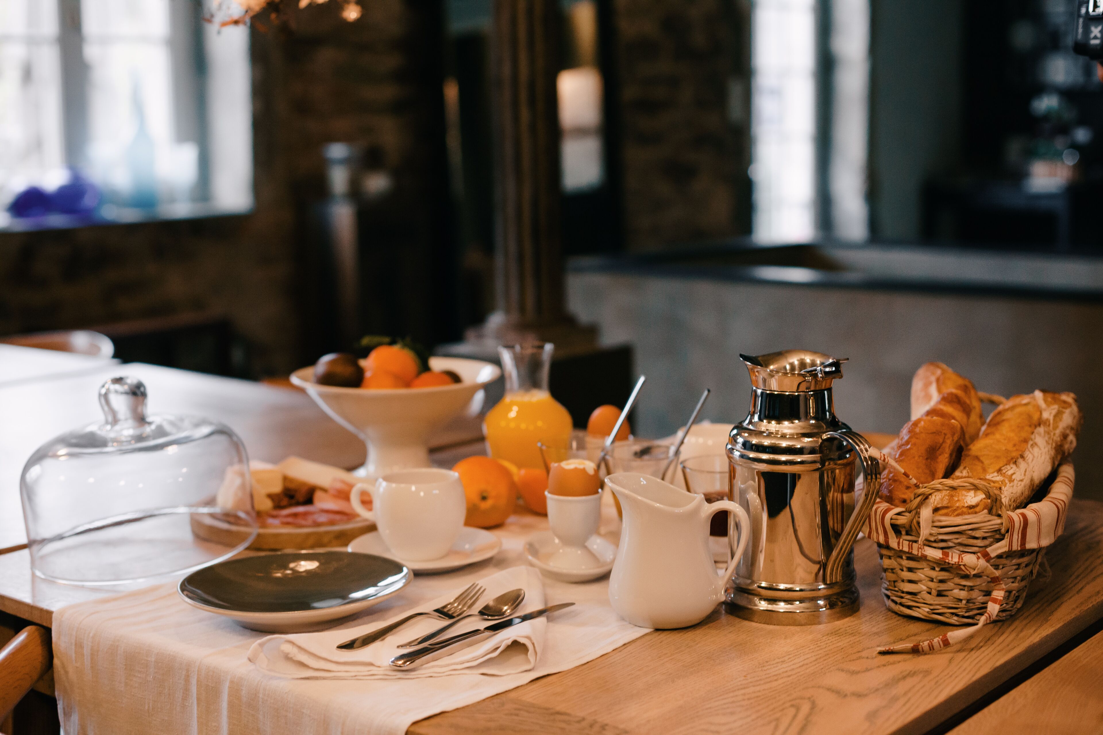 A breakfast table with a pitcher, cups, a basket of bread, and a bowl of fruit.
