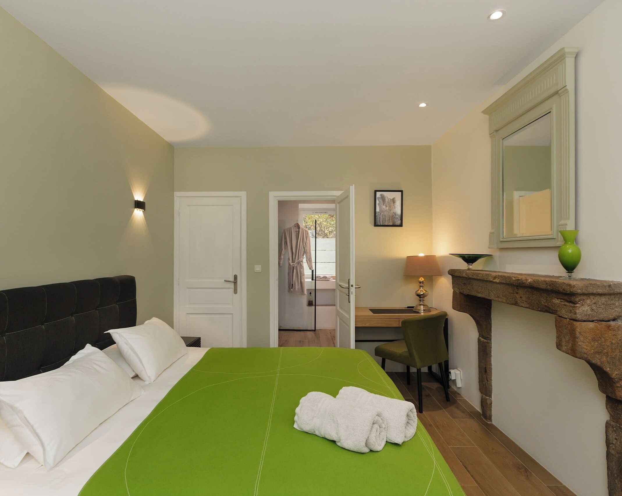 A hotel room with a green bed, white pillows, a black headboard, a green chair, a desk, a lamp, a mirror, and a stone fireplace.