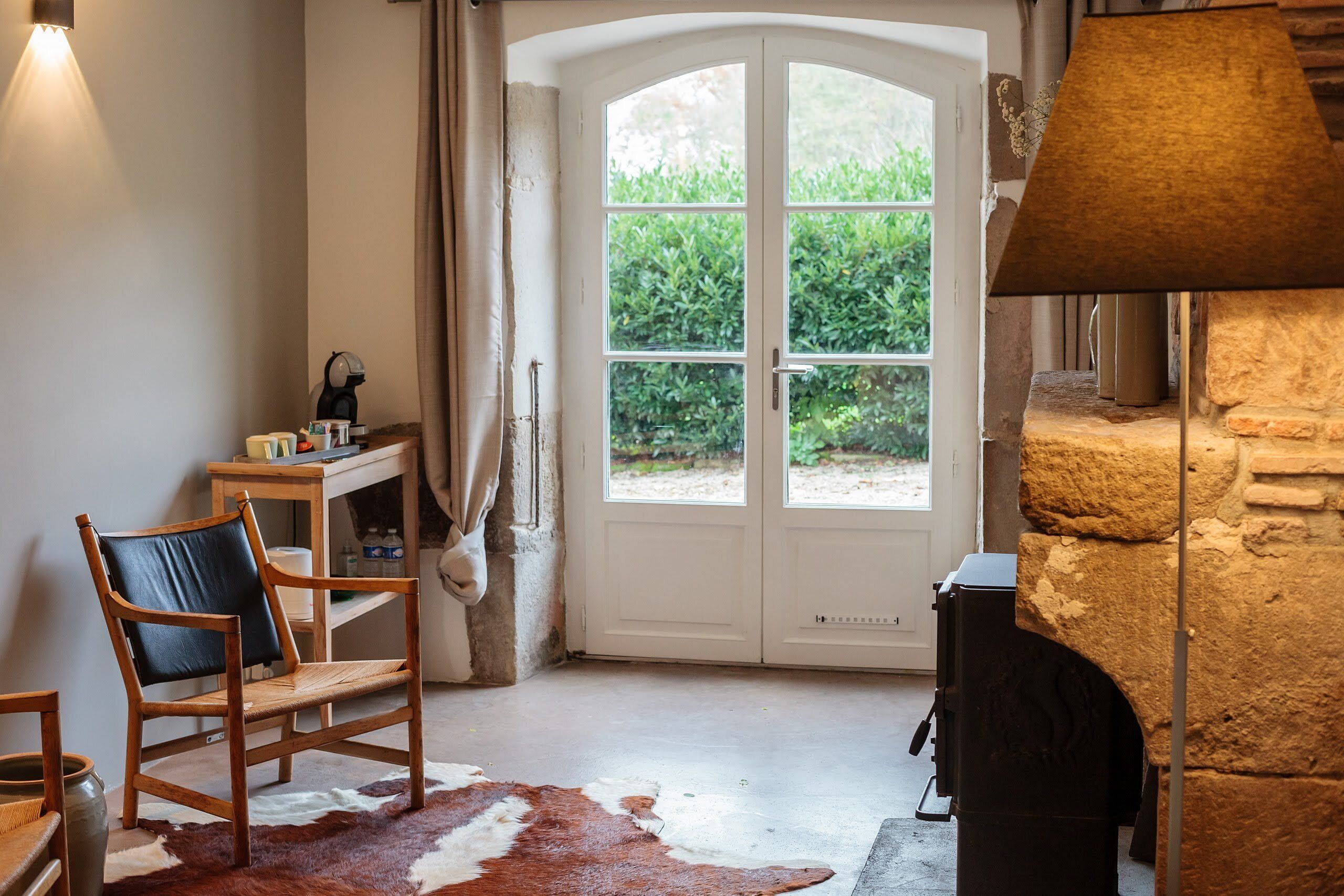 Una habitación acogedora con chimenea de piedra, una silla de madera, un tapiz de cuero de vaca y vistas a un jardín a través de una puerta blanca.
